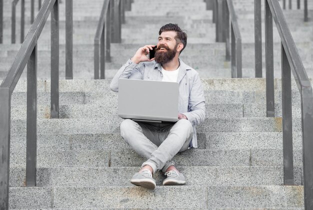 Happy man talking on mobile phone working on laptop Smiling man making mobile call on stairs