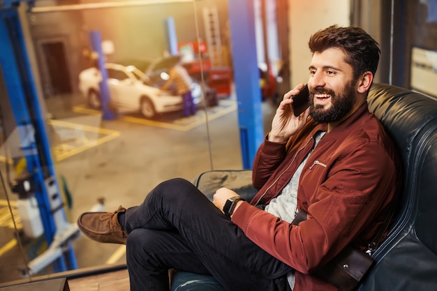 Happy man talking on smartphone, sitting on the couch, while auto technician repairing his car