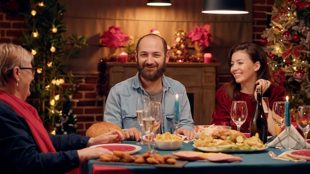 Happy man talking with senior woman and smiling at camera while\
enjoying christmas dinner. cheerful festive family members sitting\
at home while celebrating winter holiday. handheld shot