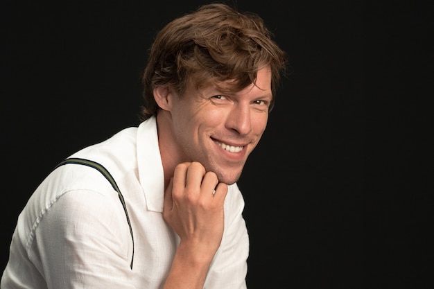 Happy man toothy smiles touching his chin by hand while looking at camera. Young smiling handsome wearing white shirt isolated on black wall.