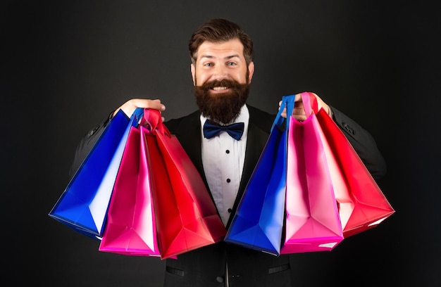 Happy man in tuxedo bow tie formalwear on black background\
holding purchase shopping bags