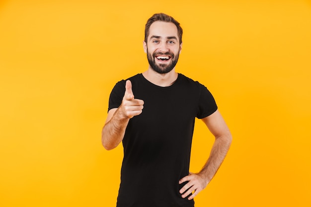 happy man wearing basic black t-shirt smiling and pointing finger at you isolated over yellow wall