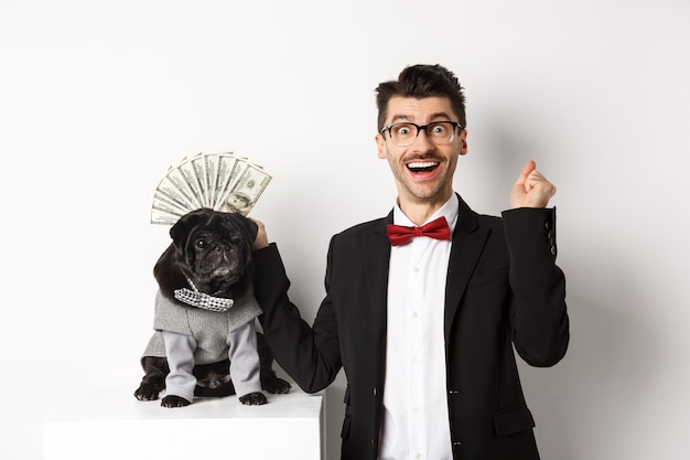 Happy man winning money, wearing costume and showing dollars near his cute black dog in suit, standing over white.
