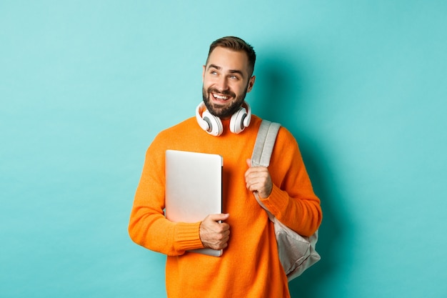 Happy man with backpack and headphones, holding laptop and smiling, looking left thoughtful, standing  