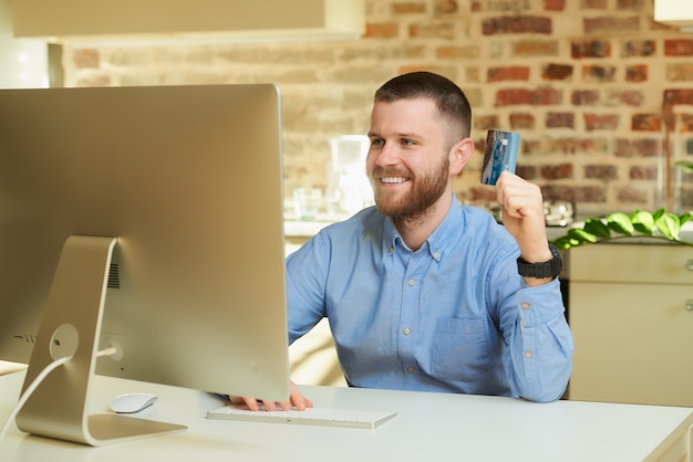 A happy man with a beard sits in front of the computer and choosing products on an online store at home