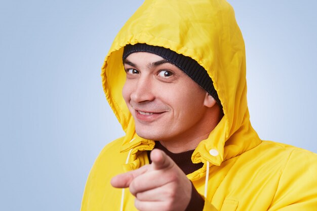 Happy man with dark appealing eyes wears yellow coat with anorak, points with fore finger directly, chooses someone, isolated over blue wall. Selective focus. Man indicates at you