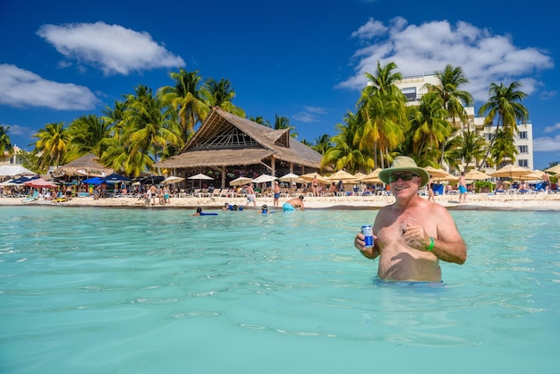 방갈로 Mujeres 섬 카리브해 Cancun Yucatan Mexico가 있는 모래 해변 근처의 청록색 물에 에너지 음료 칵테일과 쿠바 시가가 서 있는 행복한 남자