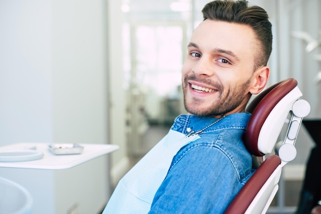 개암나무 눈과 짙은 갈색 머리를 가진 행복한 남자가 치과 의자에 앉아 치과 의사의 일에 만족하기 때문에 카메라를 똑바로 보고 웃고 있습니다