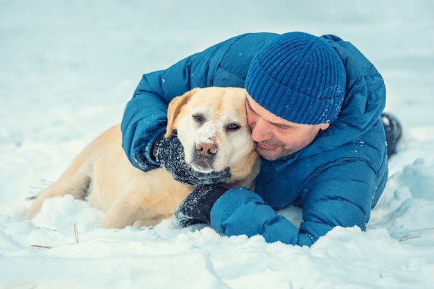 A happy man with a Labrador retriever dog lying in the snow in winter. Man hugging the dog