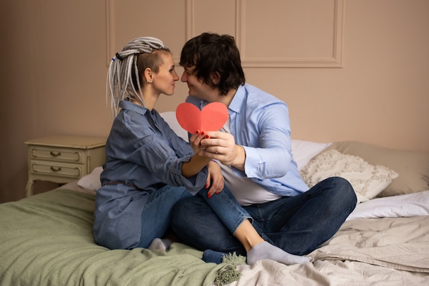 Happy man and woman as a couple at home on bed with red heart valentine in hands. Celebrating Saint Valentine's Day.