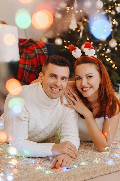 A happy married couple is lying on the floor near the Christmas tree at home.