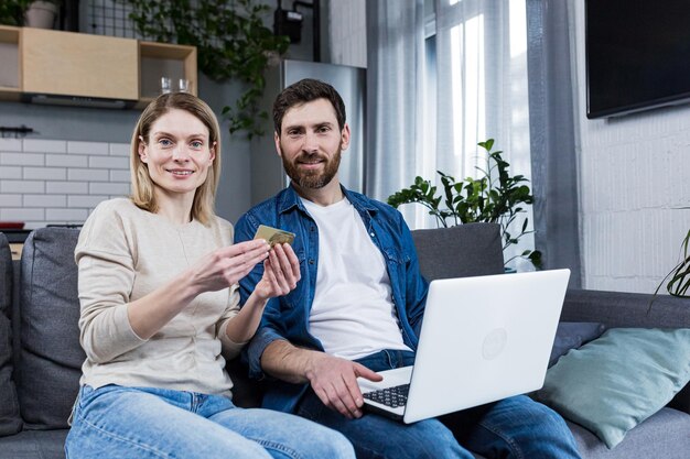 Happy married couple man and woman sitting at home on the couch using laptop for online shopping holding a credit bank card