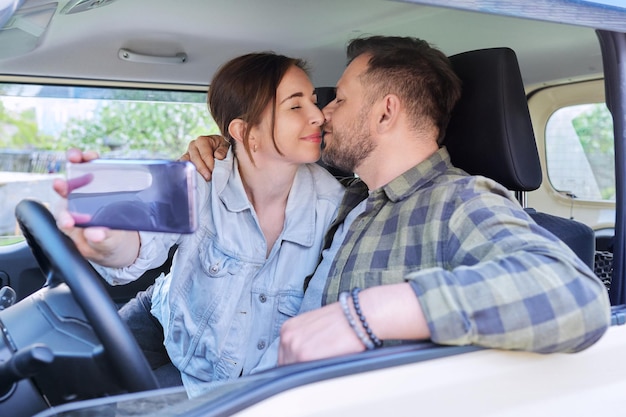 車に座っているスマートフォンの夫と妻でselfie写真を撮る幸せな夫婦