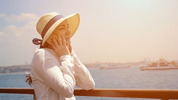 Happy married lady in elegant hat travels by ship and talks on phone standing on deck against amazing seascape closeup