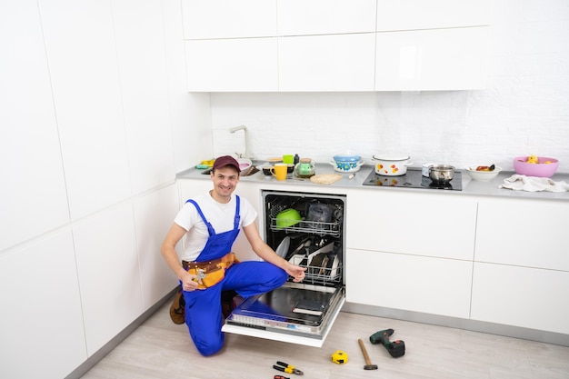 Счастливый мастер. Мужчина-техник сидит рядом с посудомоечной машиной с отверткой на кухне с инструментами и улыбается.