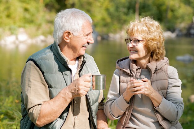 Happy mature active hikers with hot tea looking at one another while enjoying rest on sunny day