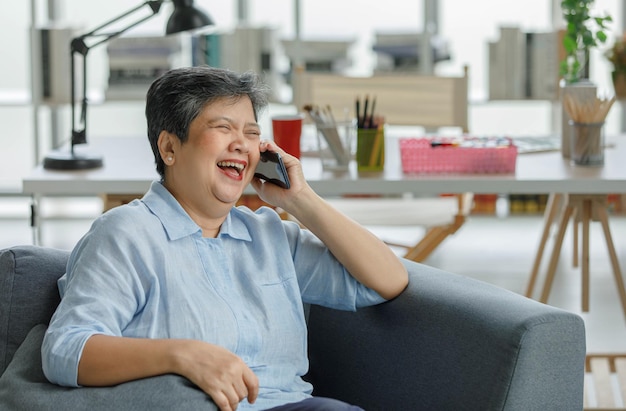 Happy mature Asian woman laughing at joke while sitting on sofa and talking on smartphone in living room at home