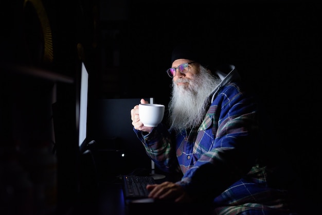 Happy mature bearded hipster man drinking coffee while working overtime at home in the dark