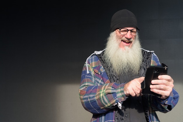 Photo happy mature bearded hipster man using phone against black wall