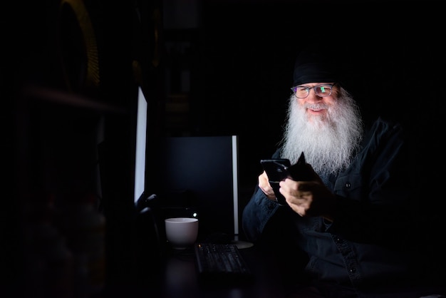 Happy mature bearded hipster man using phone while working overtime at home in the dark