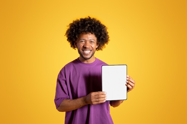 Happy mature black curly man in purple tshirt shows tablet with empty screen isolated on yellow