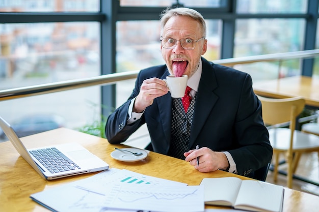 Happy mature business man laughing in spectacles in modern office. Successful senior businessman sitting in meeting room with cup of coffee. Smiling man in suit at elegant office.