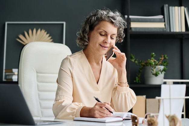 Счастливая зрелая деловая женщина с ручкой, делающая заметки и разговаривающая по телефону