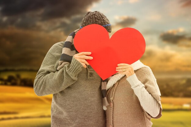 Photo happy mature couple in winter clothes holding red heart against country scene