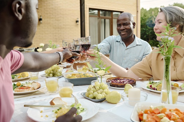 屋外ディナー中に自家製料理を提供されるテーブルのそばに座りながら、息子と妻と乾杯するワイングラスを持つ幸せな成熟した夫婦