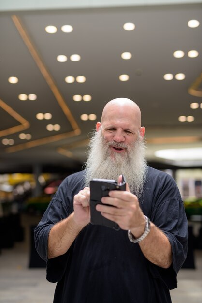 Happy mature handsome bald bearded man using phone in the city outdoors