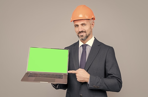 Счастливый зрелый мужчина в каске, указывающий пальцем на зеленый экран современного беспроводного ноутбука для копирования пространства, представляющего продукт
