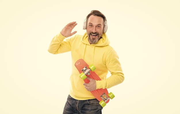 フーディでひげを生やした幸せな成熟した男のスケートボーダーは、ヘッドフォンを身に着けている音楽を聴き、楽しんで白で隔離ペニースケートボードを保持します
