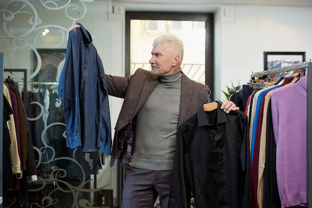 A happy mature man with gray hair and a sporty physique is choosing between two shirts in a clothing store. A male customer with a beard wears a wool suit in a boutique.