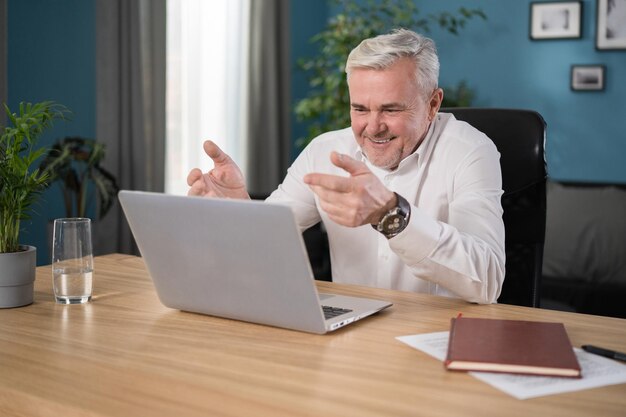 Happy mature s man sit at table at home looks at device screen use videocall application on laptop