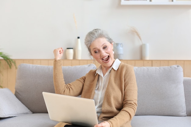 Happy mature woman celebrating online win using laptop sitting on couch at home.