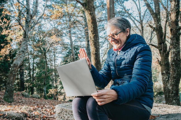 Felice donna matura che fa una chat video tramite il computer portatile durante una giornata nella foresta durante un viaggio a treck. copia spazio seduto su una panca di legno. stile di vita sano della donna anziana attiva.