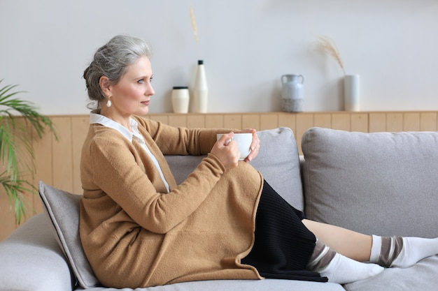 Happy mature woman resting on comfortable sofa drink coffee or tea, looking away, relaxing on cozy couch at home enjoy hot beverage