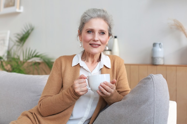 Happy mature woman resting on comfortable sofa drink coffee or tea, looking away, relaxing on cozy couch at home enjoy hot beverage