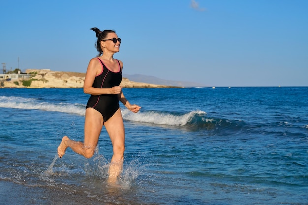 ビーチに沿って走っている幸せな成熟した女性、コピースペース。水着のジョギング、海の夕日のビーチの背景に健康的なアクティブな女性。スポーツ、ライフスタイル、休暇、中年の人々の概念