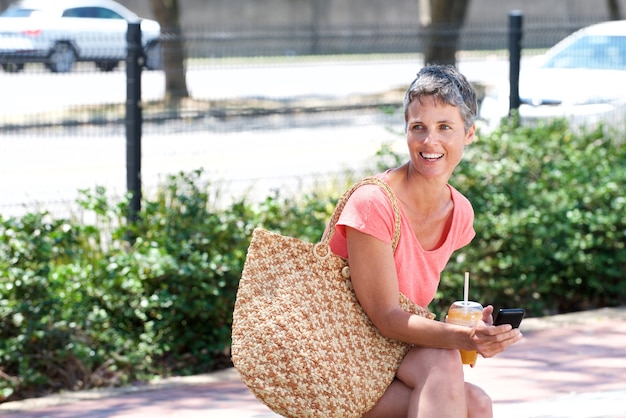 Happy mature woman sitting outdoors with drink and using phone