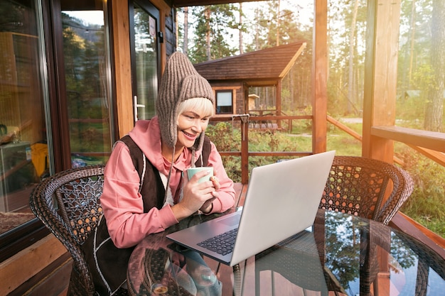 Happy mature woman sitting on terrace at sunset and using laptop while having video call.