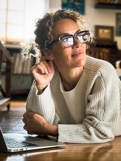 Photo happy mature woman thinking happy at home laying on the wooden floor and smiling looking outside open laptop computer in front of her modern online people planning vacation or business alone