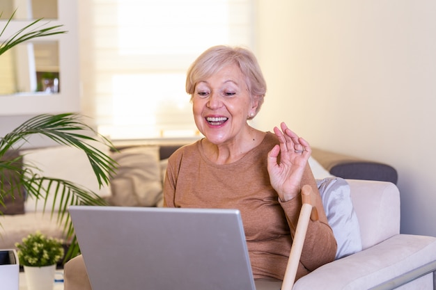 Happy mature woman waving to someone while having a video call over laptop at home. Gray-haired senior woman waving hand in front of laptop while having video call with her family members.