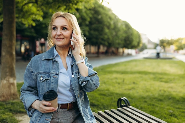 Happy mature woman wearing stylish clothes Woman resting on a bench drinking coffee in and using on the phone in the city Lifestyle concept