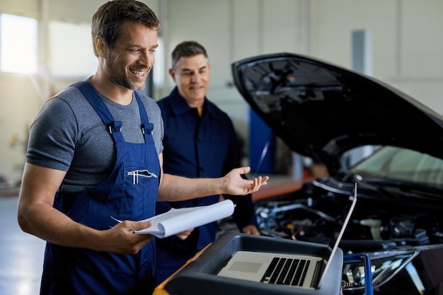 Happy mechanic using laptop while doing car diagnostic with his colleague in auto repair shop