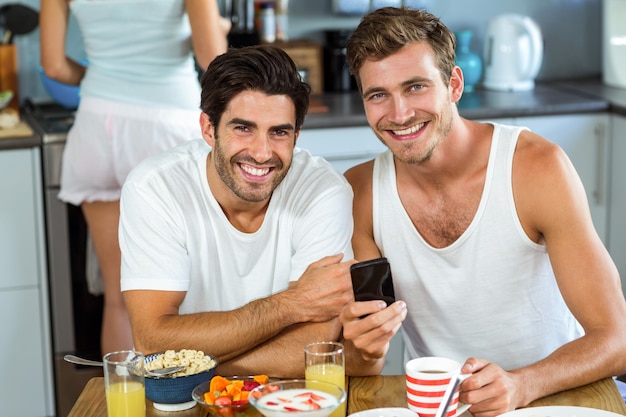 Happy men holding mobile phone while having breakfast at home