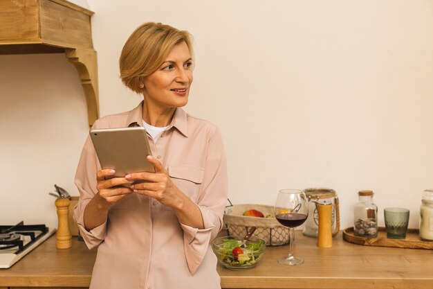 Felice di mezza età di 50 anni donna con tavoletta digitale seduto in cucina a casa. una signora anziana matura che tiene in mano un computer pad, trova la ricetta, fa acquisti online, effettua una videochiamata.