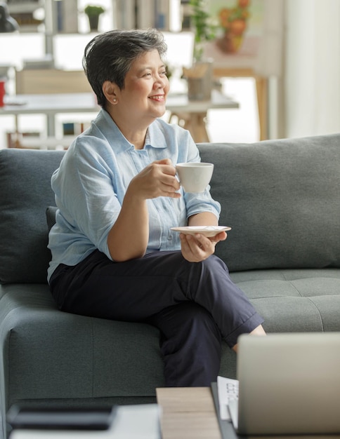 Happy middle aged Asian female smiling and looking away while sitting on sofa and enjoying tea on weekend day in modern living room