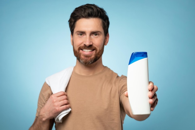 Happy middle aged bearded man showing blank shampoo bottle to camera and smiling standing over blue