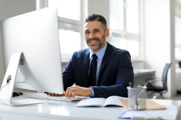 Happy middle aged businessman in suit sitting in office and smiling at camera working with modern computer free space